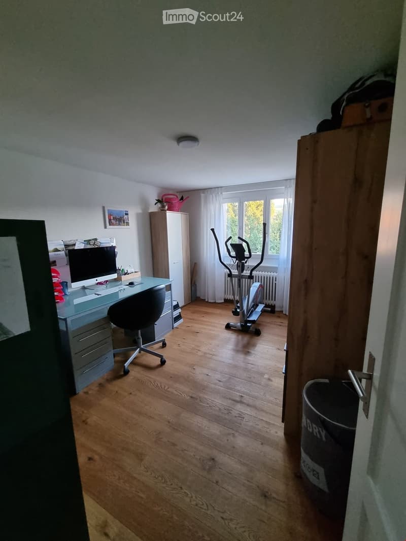 A well-lit room with wooden flooring, a desk with a computer monitor, a chair, a cabinet, an exercise bike, and a radiator near the window.