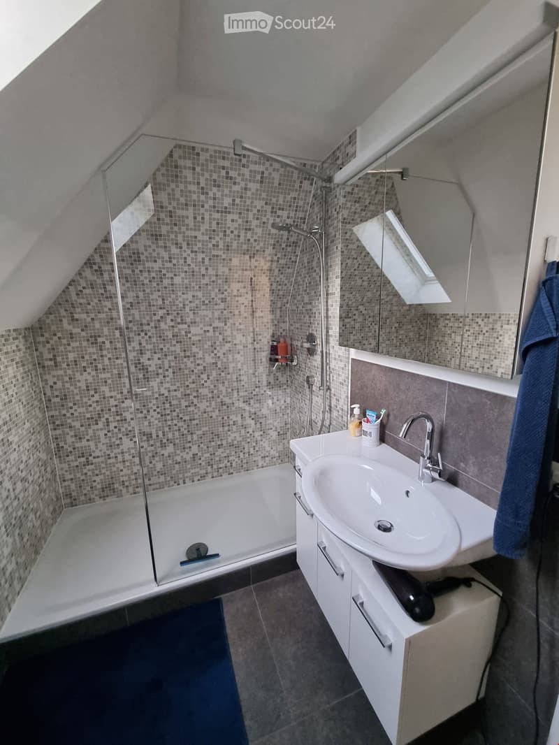Modern bathroom with a bathtub, sink, mirror, and various toiletries. The space features a sloped ceiling and a glass-enclosed shower area.