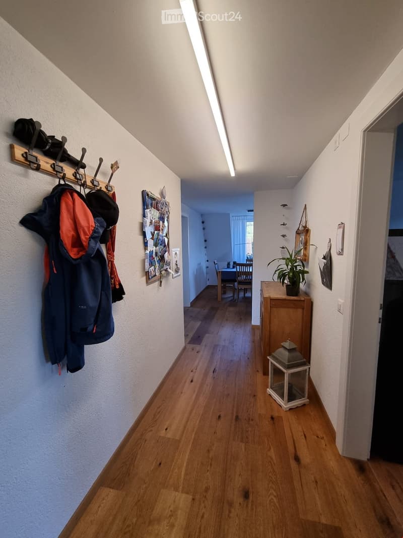 A narrow hallway with wooden flooring, white walls, and a coat rack holding jackets and hats. A small table with a plant and lantern is visible, leading to a dining area with a table and chairs.