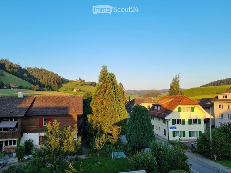A peaceful village scene with traditional houses, green fields, and trees under a clear blue sky.