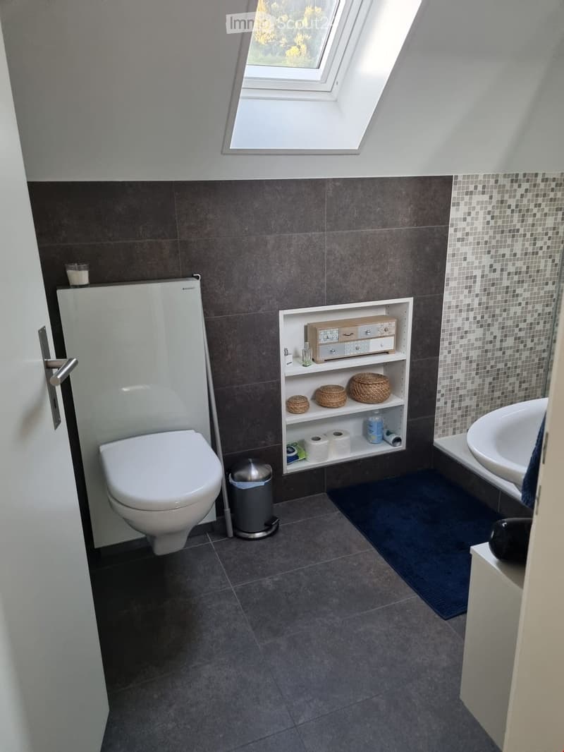 Modern bathroom with dark grey tiles, white toilet, sink, built-in shelf with toiletries, and a small window at the top.