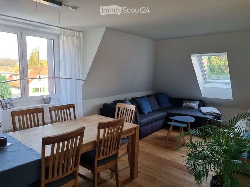 Bright living and dining area with wooden flooring, large windows, a black sectional sofa, a wooden dining table with chairs, and a potted plant.