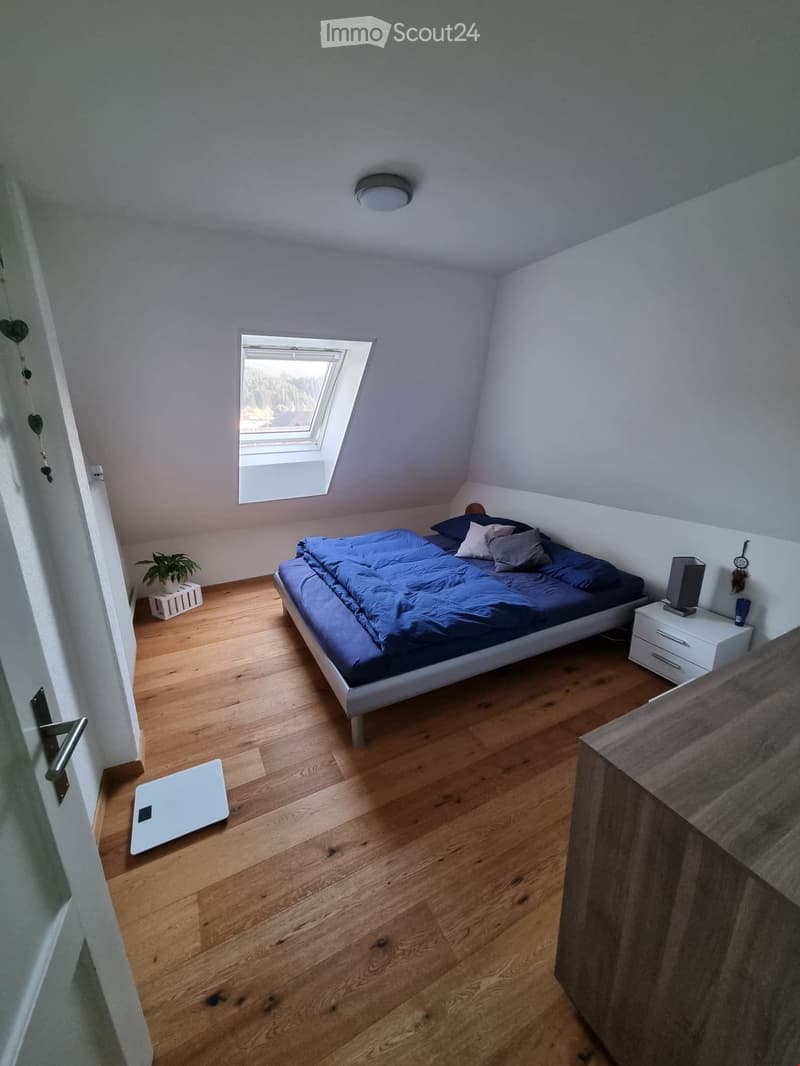 Cozy bedroom with wooden flooring, a bed with blue bedding, a nightstand with a lamp, a small plant, and a scale near the door.
