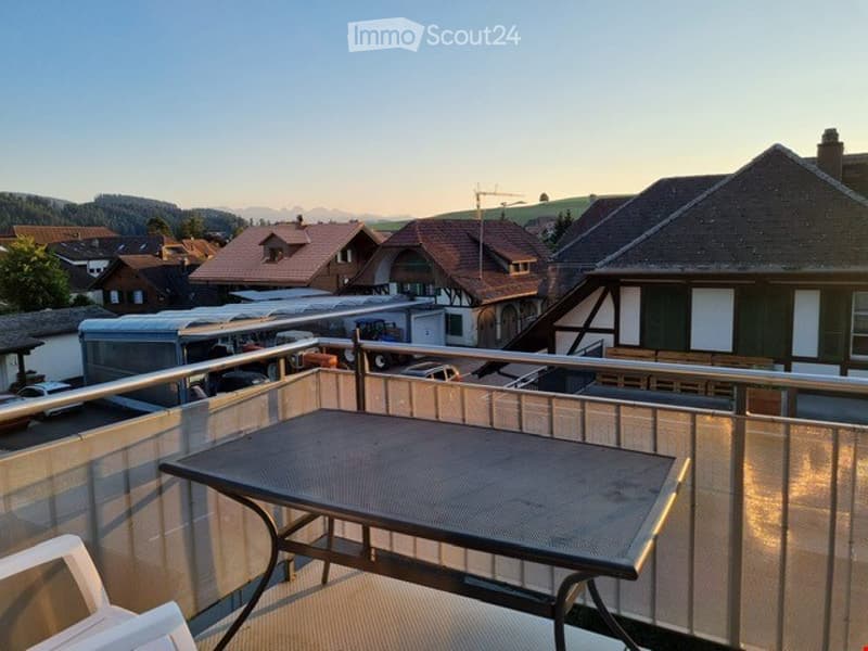 Balcony with a table and chairs, overlooking a scenic town with houses and mountains in the background.