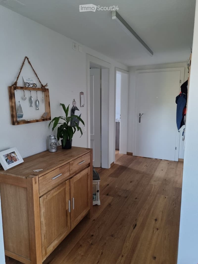 Hallway with wooden flooring, white walls, and several doors leading to other rooms. A wooden sideboard with a picture frame, a potted plant, and decorative items is visible on the left side.