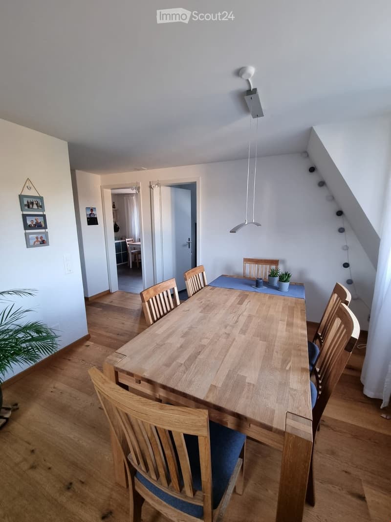 Dining area with a long wooden dining table surrounded by wooden chairs, a small potted plant on the table, and a blue runner. The room has wooden flooring and white walls, with a hallway leading to another room visible in the background.
