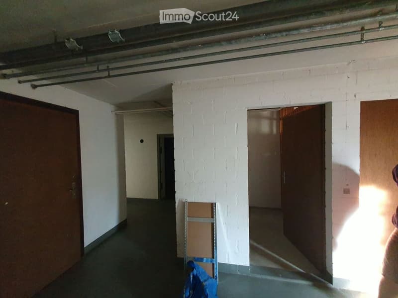 Empty hallway with white brick walls, wooden doors, and exposed pipes on the ceiling. Natural light enters from the right side.