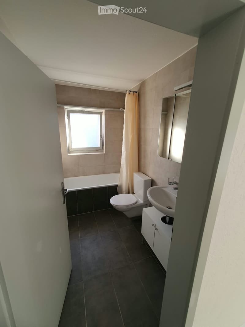 Modern bathroom with dark tiled floor, white bathtub, toilet, sink, and mirror. Features a window with a curtain and a small cabinet under the sink.