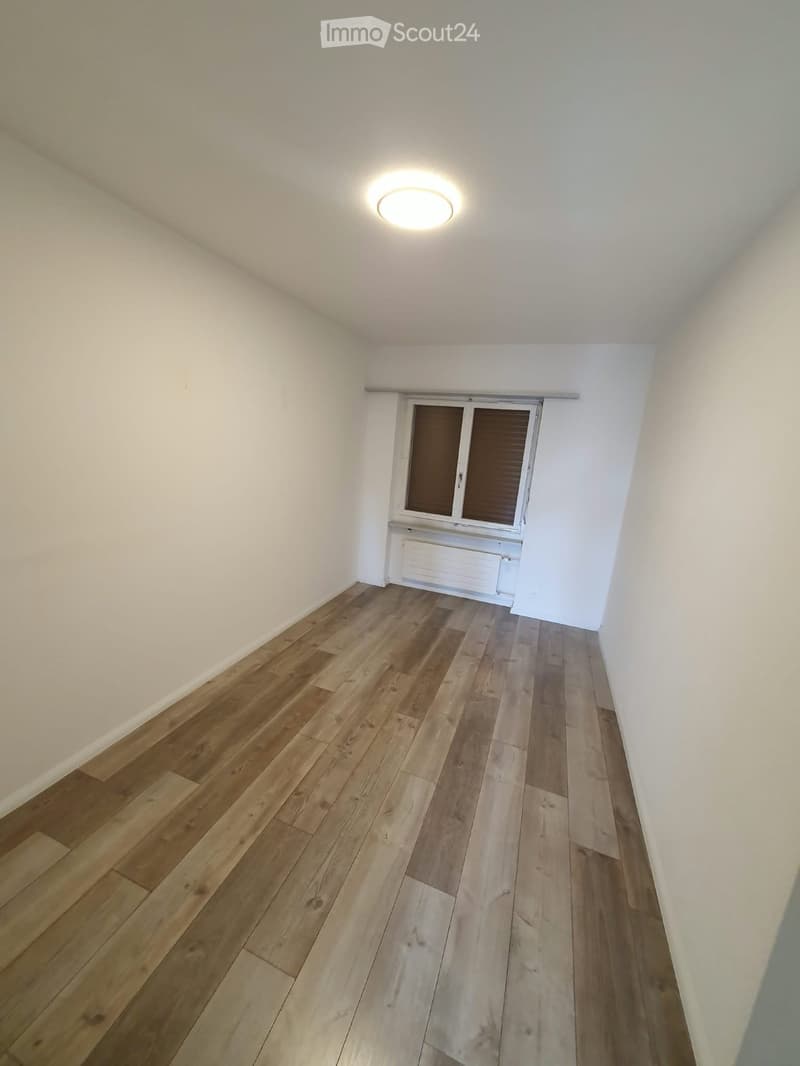 Empty room with wooden flooring, white walls, a window with blinds, and a ceiling light fixture.