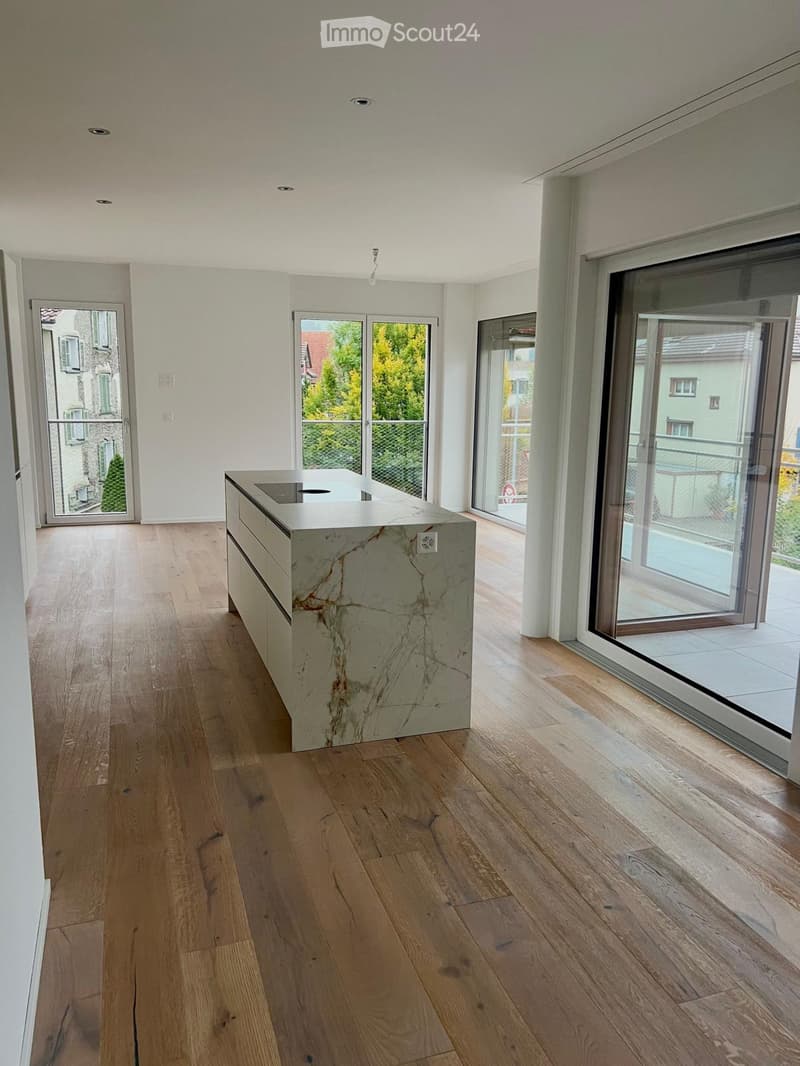 Modern, minimalist kitchen with light wooden flooring, a central marble kitchen island, and large glass doors leading to a balcony.