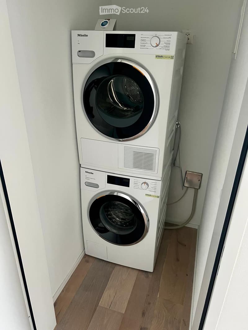 A compact bathroom with a white washing machine and dryer stacked vertically, light wooden flooring, and white walls.