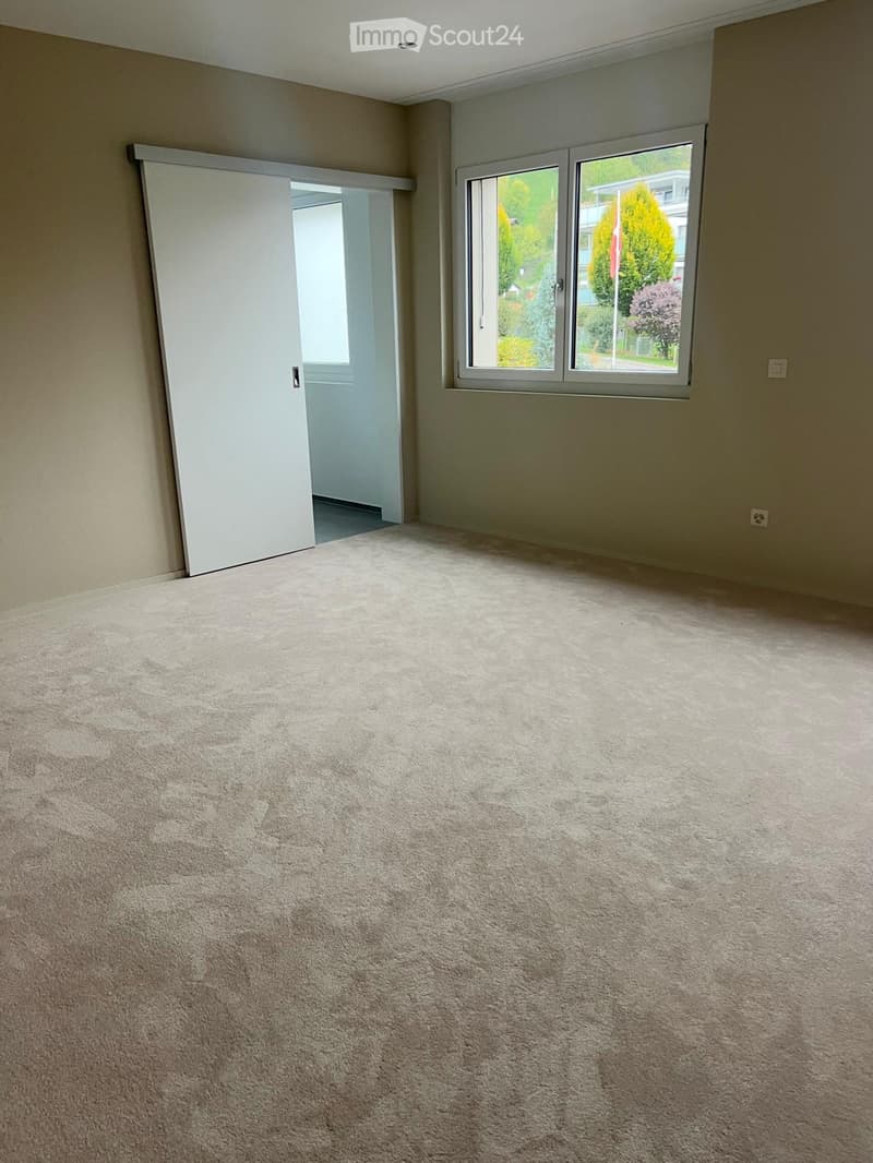 Empty room with beige carpet, white walls, large window with view of trees, and a sliding door leading to another space.
