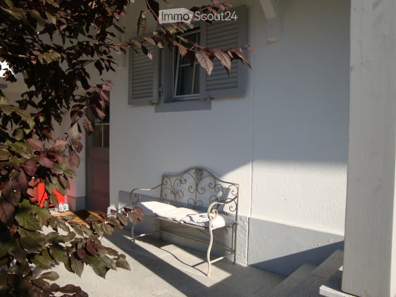 A small outdoor seating area with a white metal bench featuring ornate scrollwork and a cushioned seat, placed on a tiled patio next to a white building with shutters and a window.