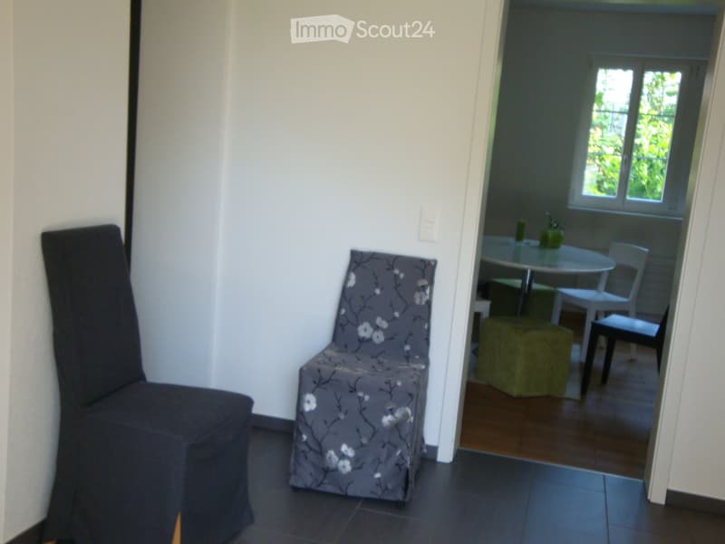 A small, furnished room with a grey chair, a floral-patterned chair, a small round table, and a green stool visible in the adjacent space.