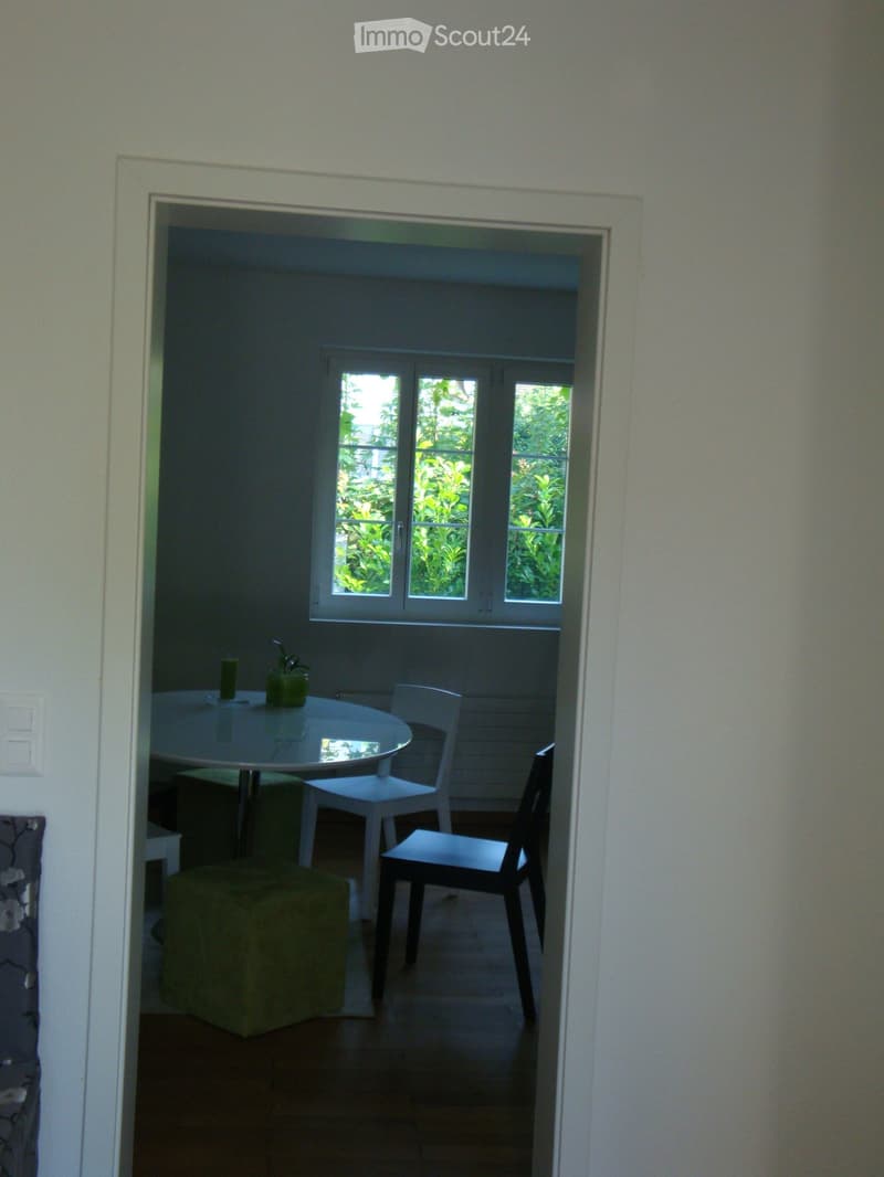 A small, furnished room with a round table, two chairs, a stool, a candle, and a small plant. The room has large windows with a view of greenery outside.