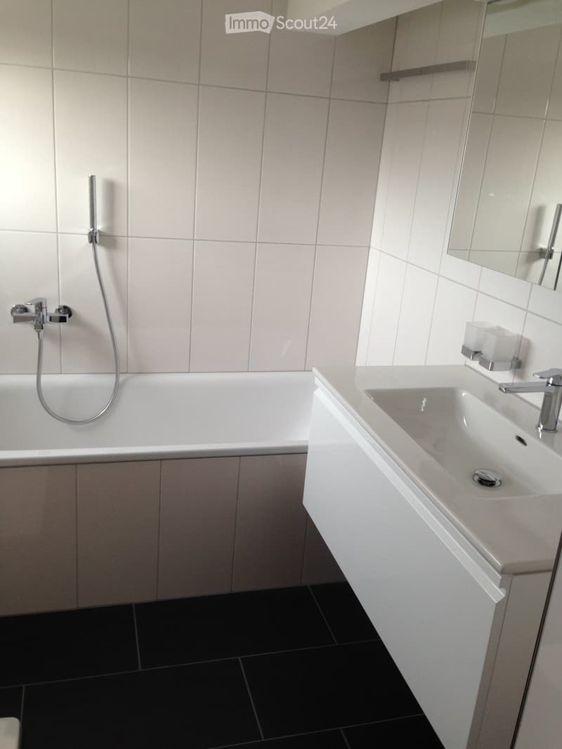 Modern bathroom with white tiles, a bathtub, a sink, and a mirror. The floor is black tiled.