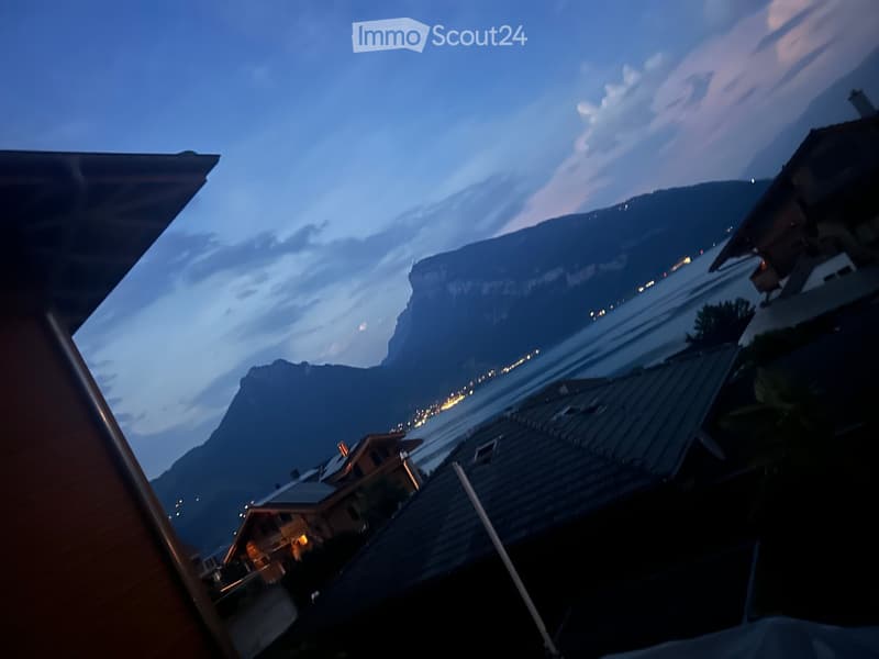 Balcony overlooking a scenic view of a lake and mountains at dusk, with distant lights from buildings and a calm water surface.