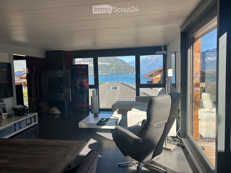 Modern living room with large windows offering a scenic mountain and lake view, featuring a white leather sofa, coffee table, fireplace, and a sleek chair.