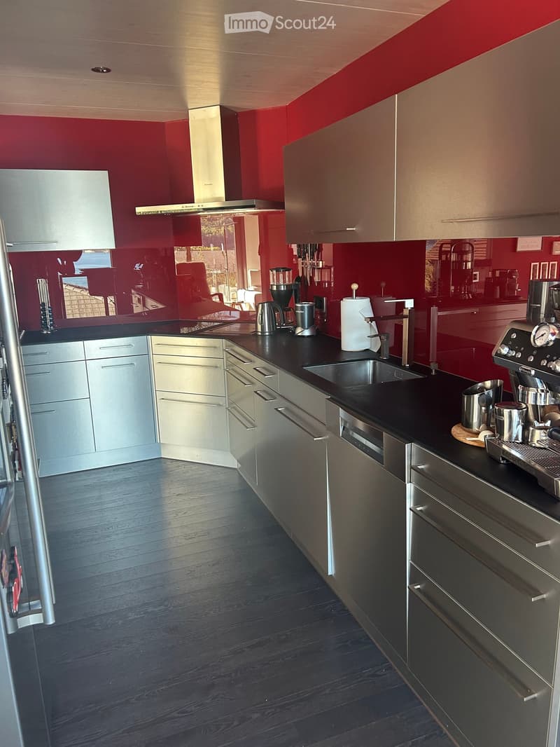 Modern kitchen with dark wooden flooring, stainless steel appliances, and red accent walls. Features include a refrigerator, cabinets, sink, coffee maker, blender, and oven.