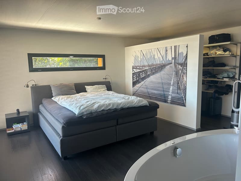 Modern bedroom with a large bed, nightstand, shelves, and a large mirror with a cityscape print. The room features dark wooden flooring and a bathtub in the foreground.