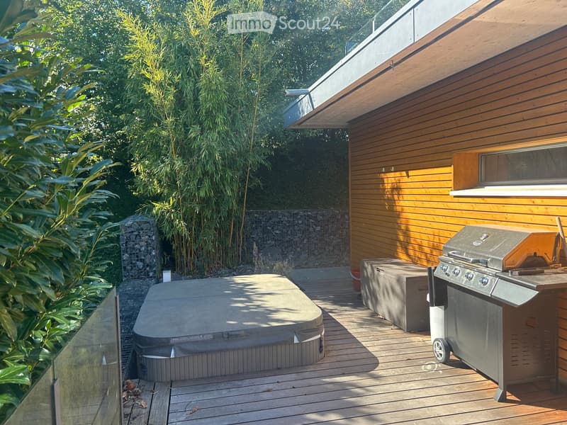 Wooden deck with a hot tub, barbecue, and storage bench, surrounded by bamboo plants and a stone wall.