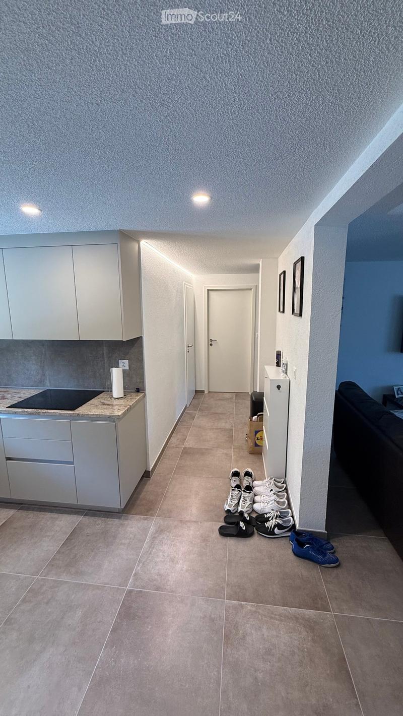 A clean, modern hallway with tiled flooring, white walls, and recessed lighting. A small kitchen area is visible on the left, and several pairs of shoes are neatly arranged on the floor near the entrance.