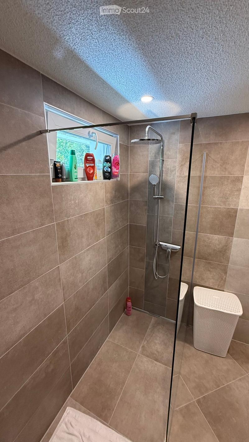 Modern bathroom with a glass-enclosed shower, tiled walls and floor, and a window with a shelf holding various bottles.