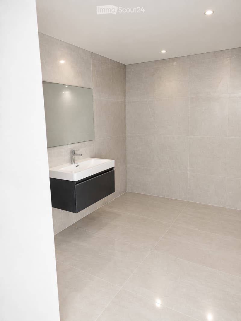 Modern bathroom with beige marble tiles, a white sink, a large mirror, and a black cabinet.