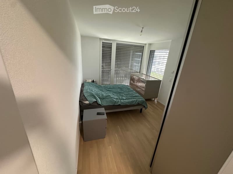 Bedroom with wooden flooring, a bed with a green duvet, a nightstand with a remote control, and a baby crib near the window.