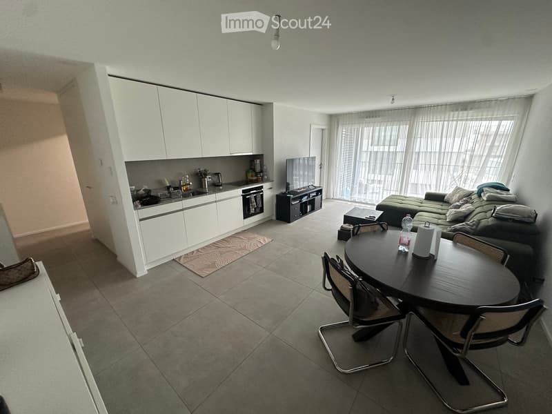 Open-plan living and dining area with a sofa, coffee table, dining table with chairs, and a modern kitchen featuring white cabinets, an oven, and a refrigerator. Large windows with curtains provide natural light.