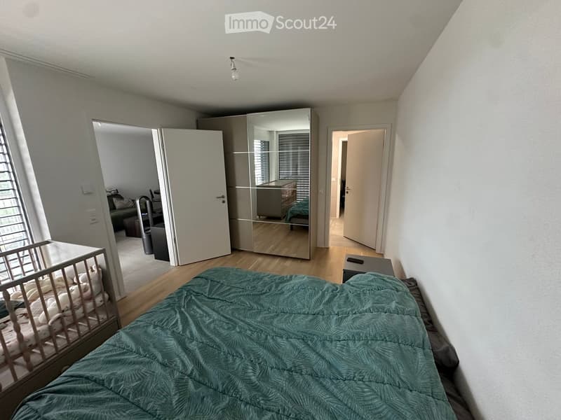 A modern bedroom with a large bed covered in a teal duvet, a full-length mirror, a wardrobe, and a small table near the door.