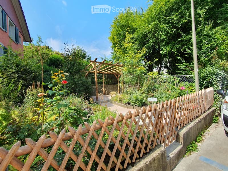 A lush garden with a wooden pergola, climbing plants, and a decorative wooden fence. The garden is enclosed by a low wooden fence and features various flowering plants and shrubs.