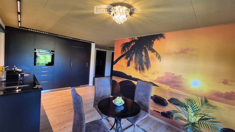 Modern kitchen with a dining area, featuring a black countertop, built-in cabinets, a stove, a refrigerator, a chandelier, and a round dining table with chairs. A large mural of a tropical beach decorates one wall.