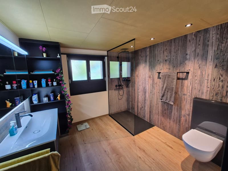 Modern bathroom with wooden flooring, a large glass-enclosed shower, a white sink, a toilet, and a black shelving unit with toiletries and decorative items.