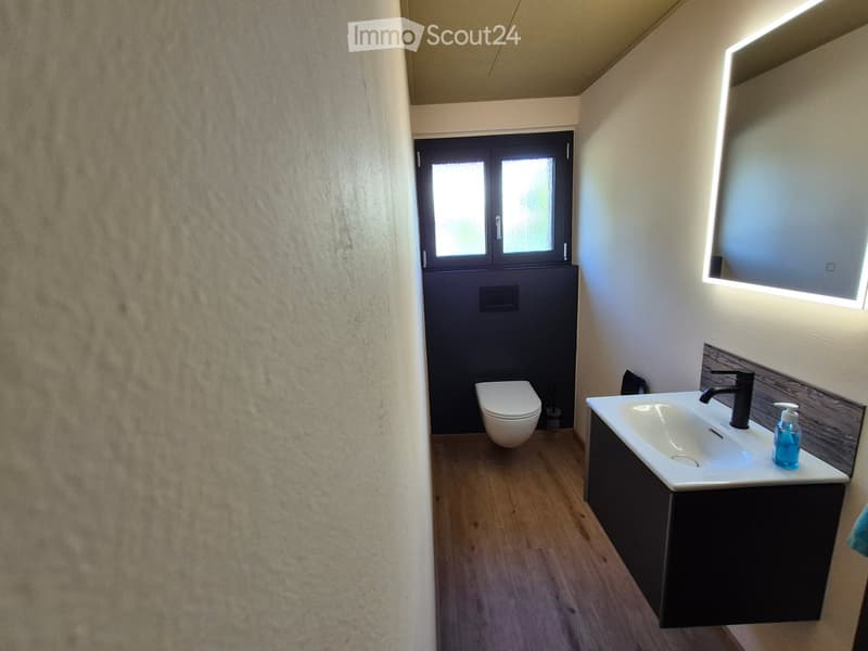 Modern bathroom with a white sink, black faucet, toilet, and a large mirror with built-in lighting. The walls are textured, and the floor is wooden.