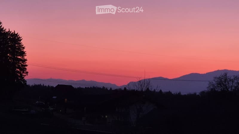 A serene view of a residential area at sunset, with silhouettes of trees, houses, and distant mountains against a pink sky.