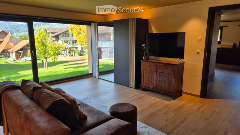 Modern living room with large windows offering a scenic view, a brown leather sofa, a wooden coffee table, a TV stand with a flat-screen TV, and a flower vase on the stand.
