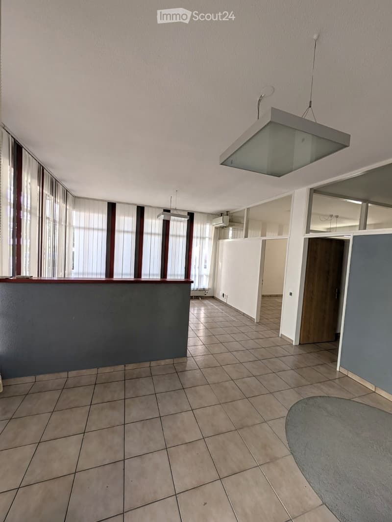 Empty room with tiled floor, large windows with blinds, grey reception desk, and a hanging light fixture.