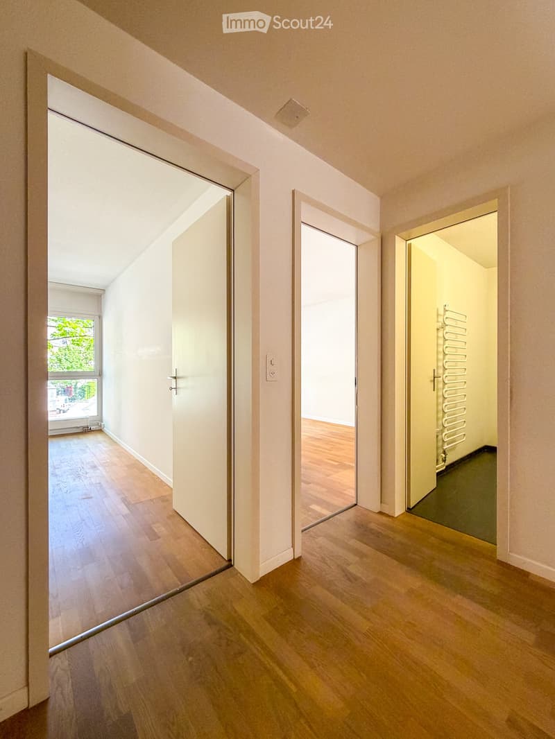 Bright hallway with wooden flooring, white walls, and multiple open doors leading to different rooms.