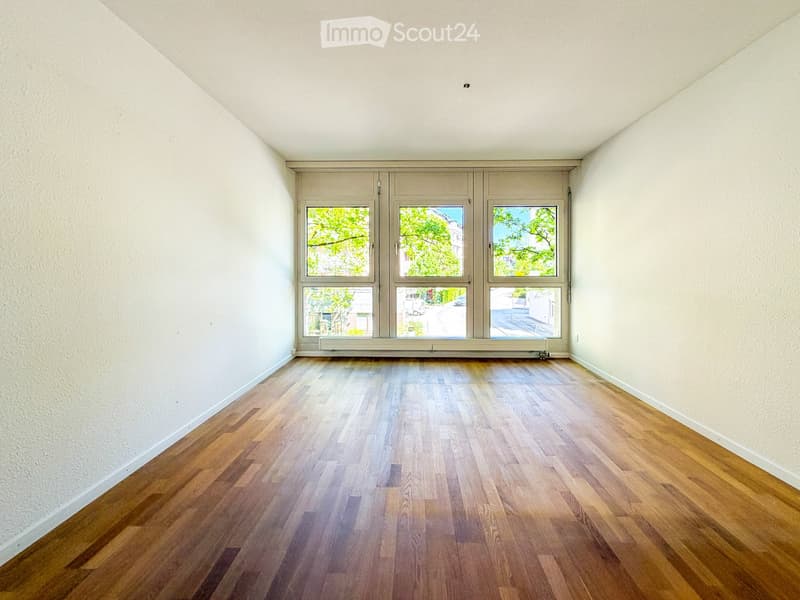 Empty room with wooden flooring and large windows offering a view of greenery and buildings outside.