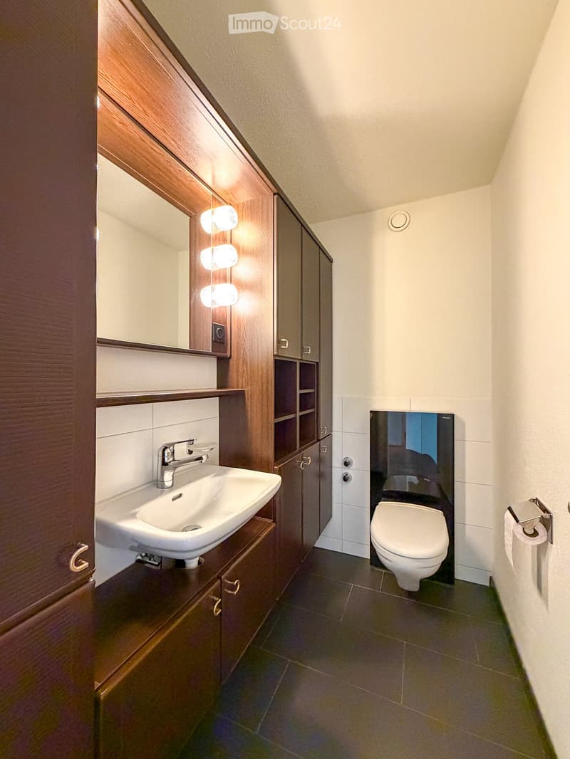 Modern bathroom with a white sink, chrome faucet, wooden cabinet, large mirror, and a toilet. The walls are tiled, and the floor is dark tiled.