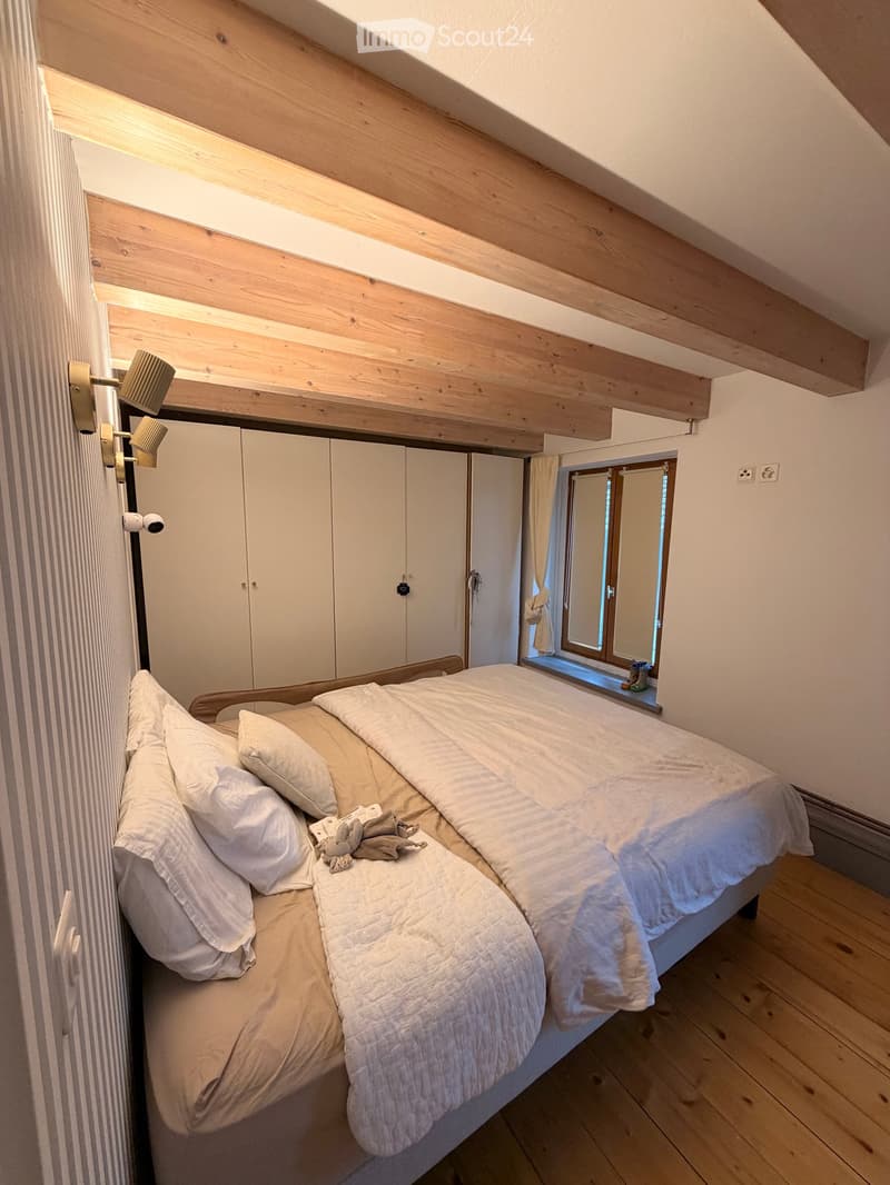 Cozy attic bedroom with wooden slanted ceilings, a large bed with white bedding, a wardrobe, and a wall-mounted lamp. The room features wooden flooring and a small window with a view.