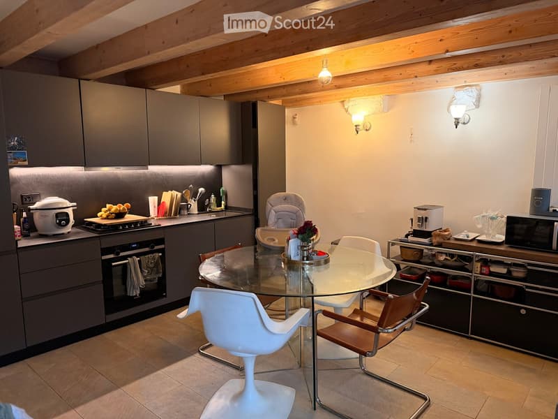 Modern kitchen with grey cabinets, stainless steel appliances, a glass dining table with white and brown chairs, and a cozy wooden ceiling.