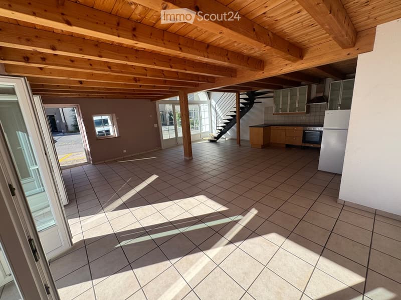 Empty room with tiled floor, wooden ceiling beams, and large windows allowing natural light. Kitchen area visible in the background with cabinets and appliances.