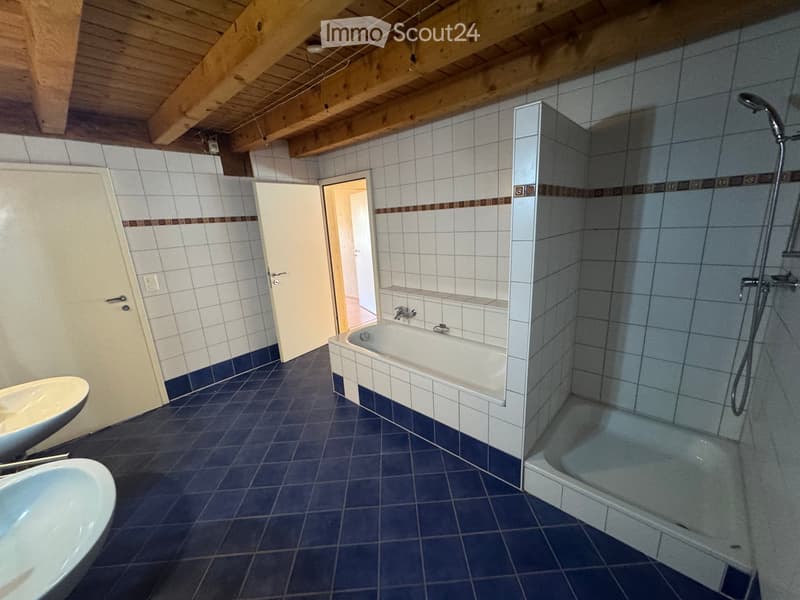 Bathroom with a bathtub, sink, and shower area. The room features white tiled walls and a blue tiled floor, with a wooden ceiling and an open doorway leading to another room.