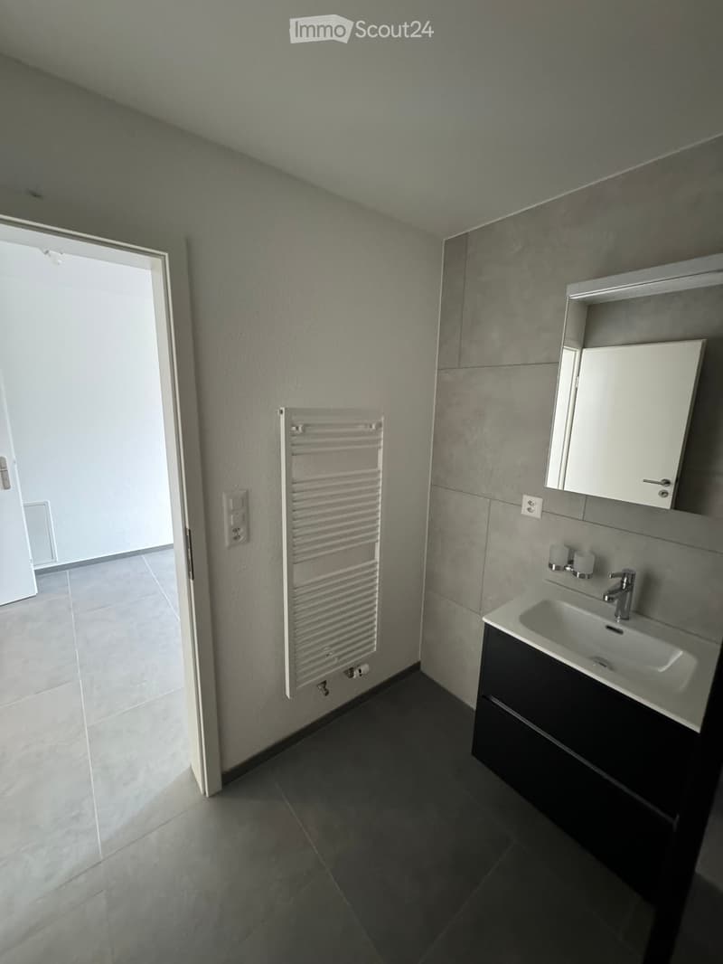 Modern bathroom with grey tiled walls and floor, white sink with dark cabinet, radiator on wall, and open doorway leading to another room.