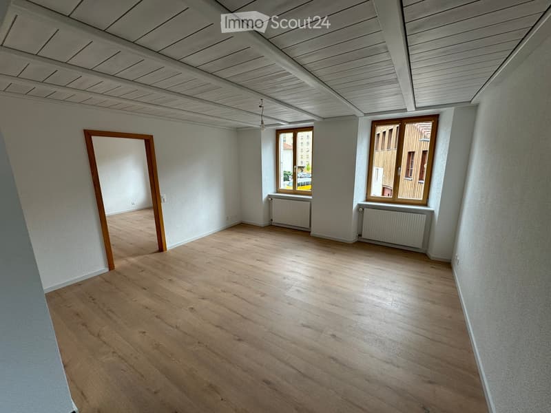 Empty room with wooden flooring, white walls, and large windows with wooden frames. Radiators are visible beneath the windows.