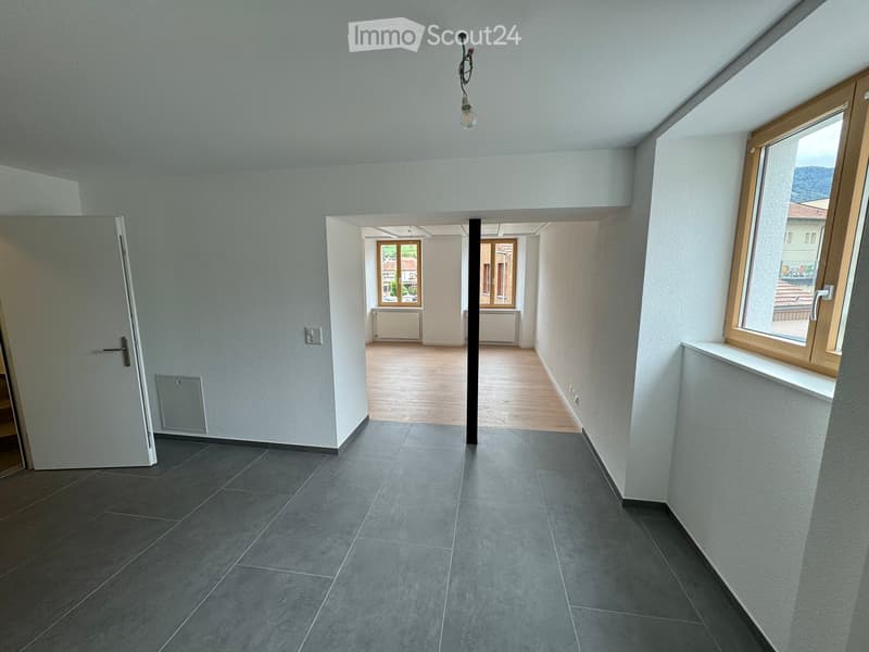 Empty hallway with grey tiled floor, white walls, and wooden door frame. Natural light from windows at the end of the corridor.