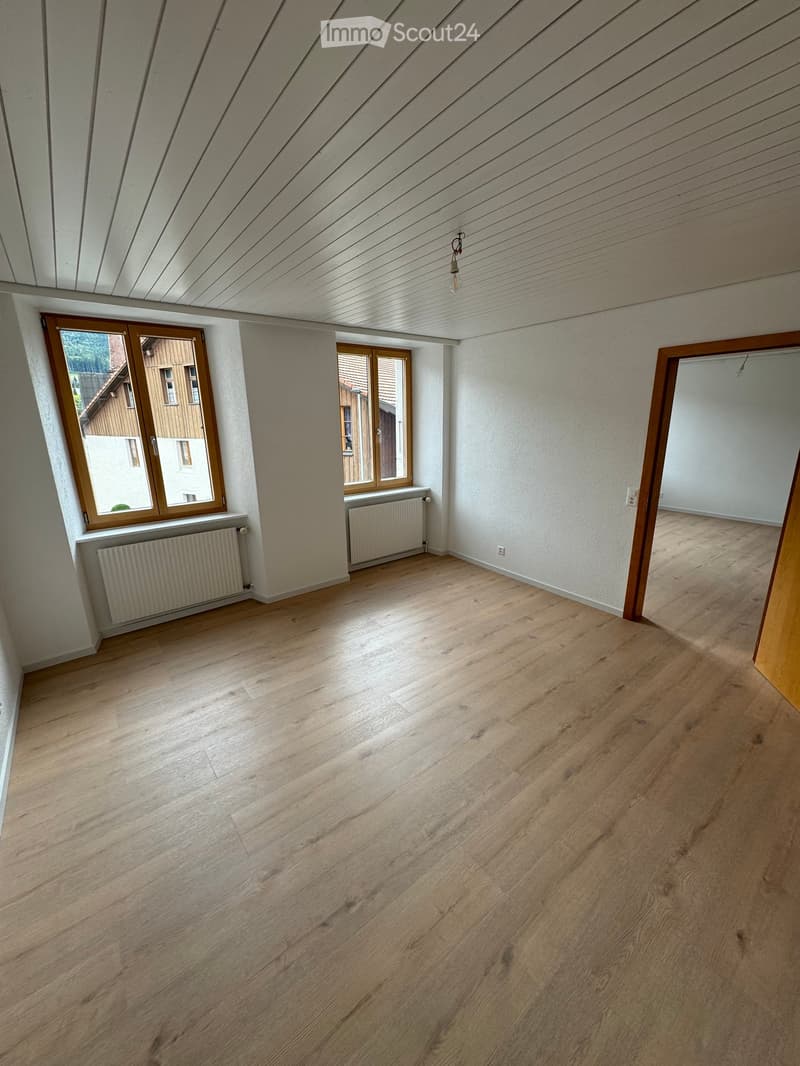 Empty room with wooden flooring, white walls, and large windows with wooden frames. Radiators are visible beneath the windows.