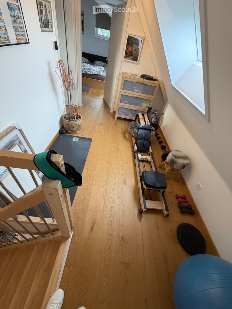 A narrow hallway with wooden flooring, leading to a bedroom area. Visible items include a yoga mat, exercise bike, small cabinet, potted plant, and framed pictures on the walls.